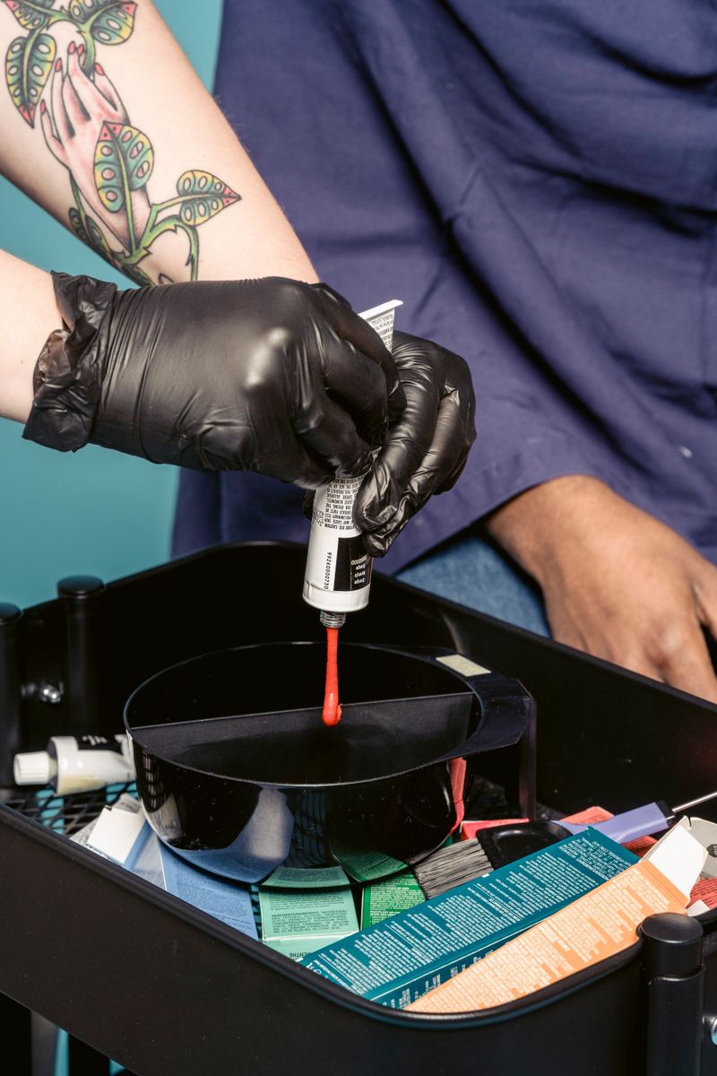 Crop woman in gloves squeezing bright dye into bowl near anonymous customer in salon